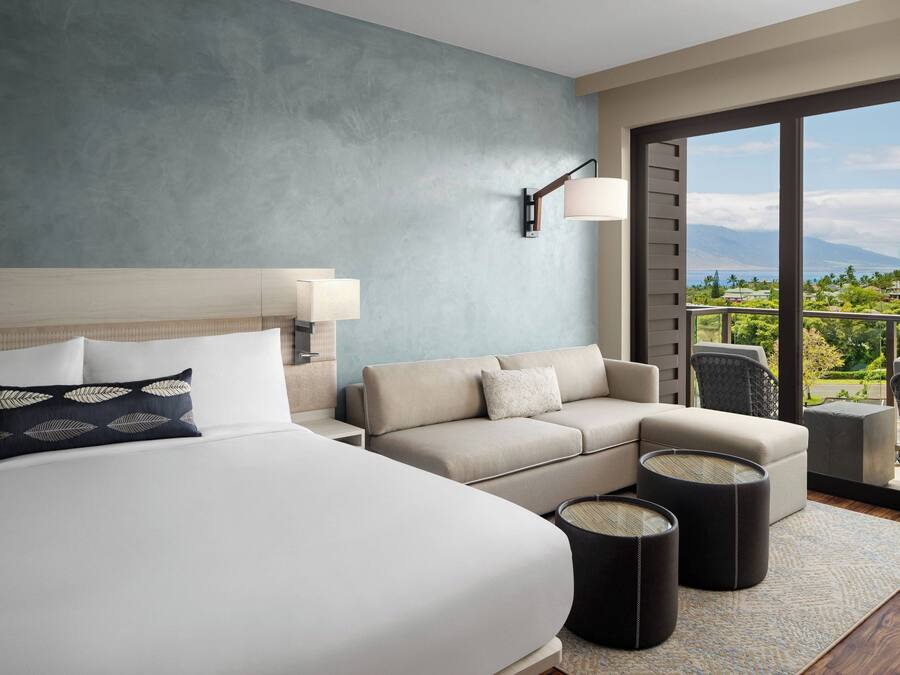A hotel room with a large bed, a sofa, a small table, and a view of a balcony with outdoor scenery.