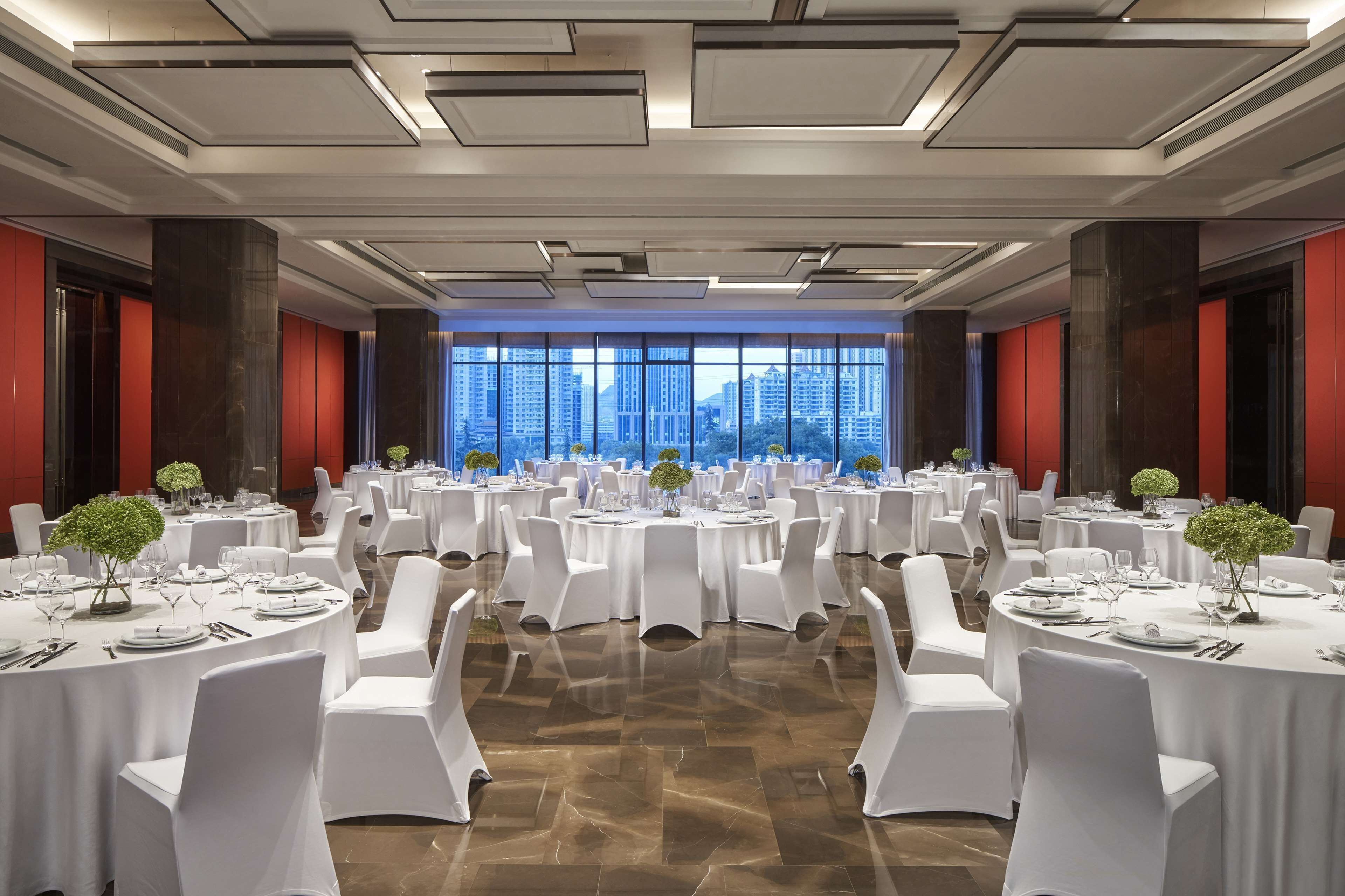 A spacious banquet hall with round tables, white chairs, and large windows offering a city view.