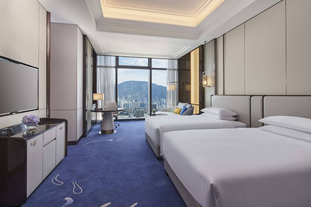 A hotel room with two beds, a desk with a lamp, a TV, and a city view through large windows.