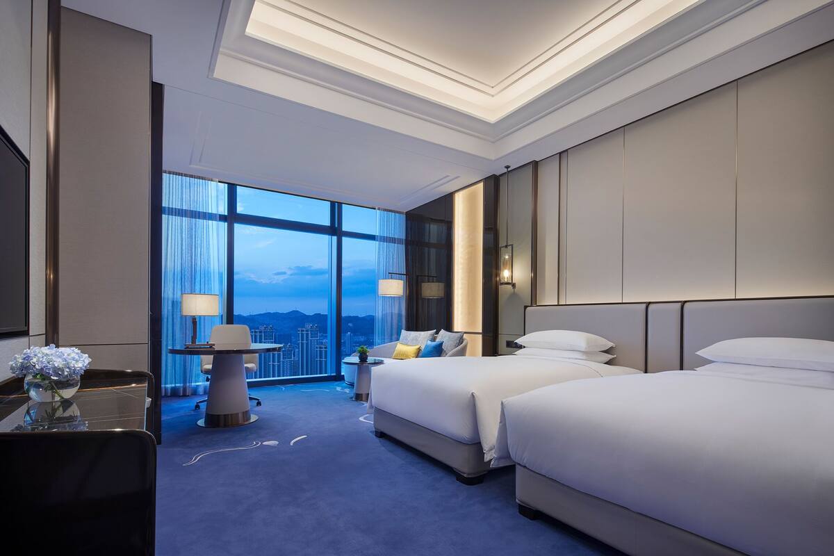A hotel room with two beds, a desk with a lamp, a chair, and a view of the cityscape through large windows.