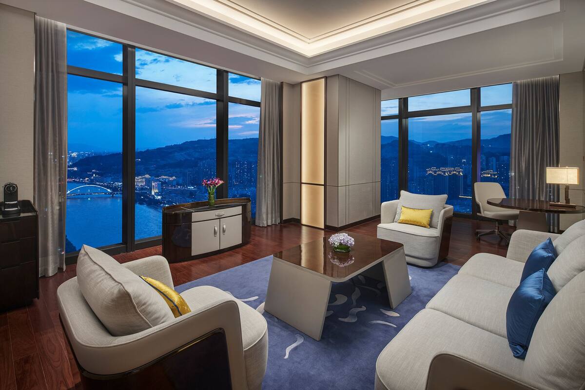 A modern hotel room with a large window offering a city view, a sofa set, a coffee table, and a desk with a lamp.