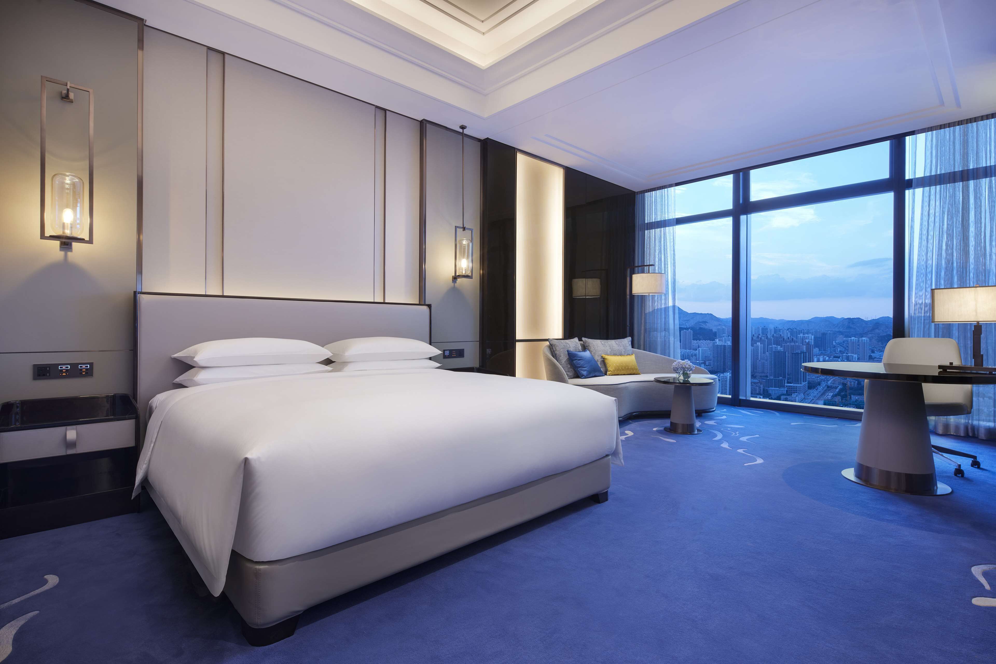 A modern hotel room with a large bed, a sofa, a desk, and a city view through large windows.