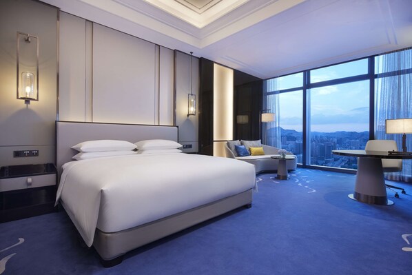 Quarto de hotel moderno com uma cama grande, um sofá, uma escrivaninha e vista para a cidade através de amplas janelas.