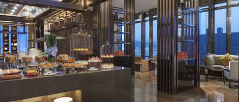 A modern hotel lobby with a buffet area, high ceilings, and large windows offering a city view.