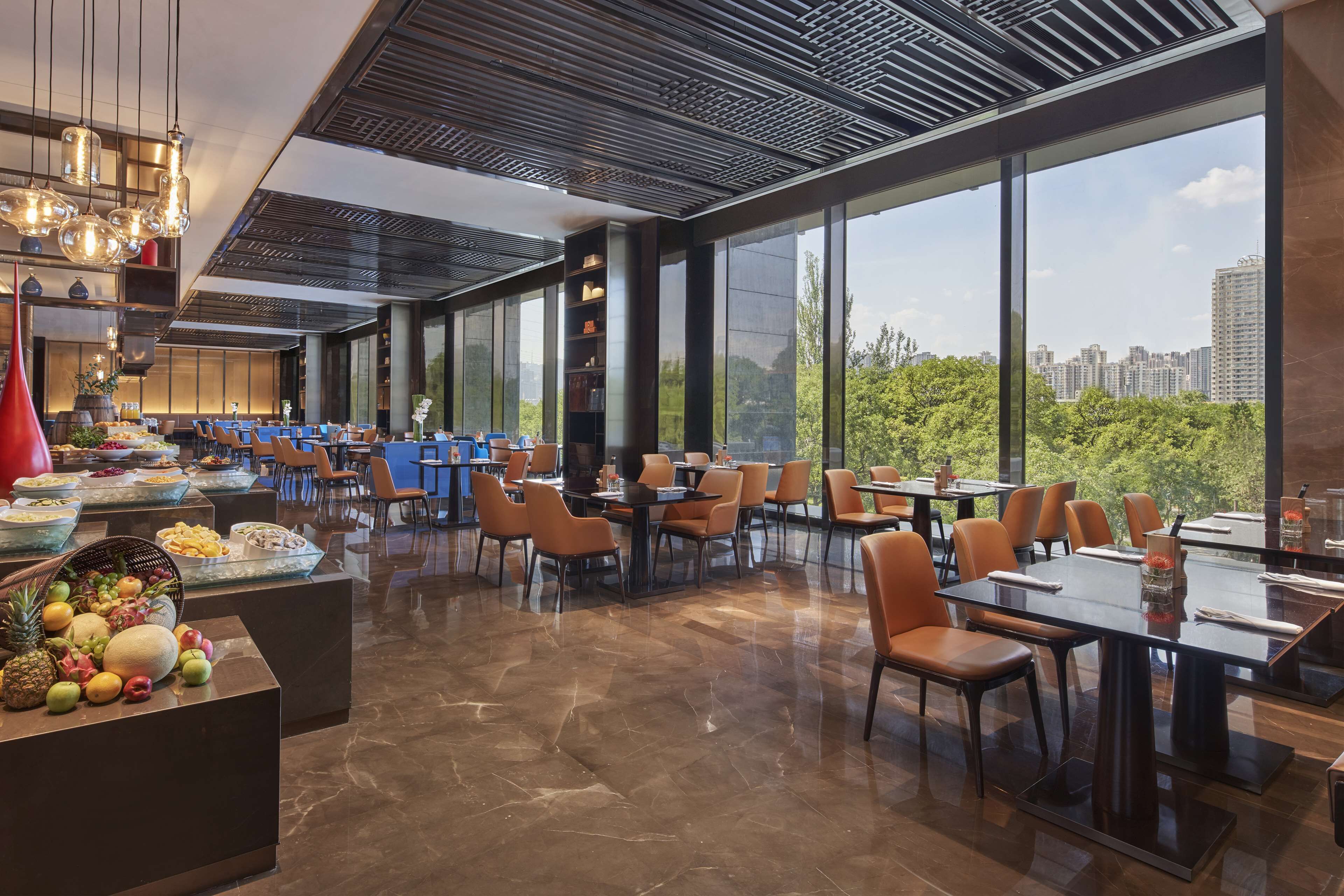 Een modern restaurant met uitzicht op de skyline van de stad, met een lange toonbank met fruit, meerdere tafels en een hoog plafond met een gedessineerd patroon.