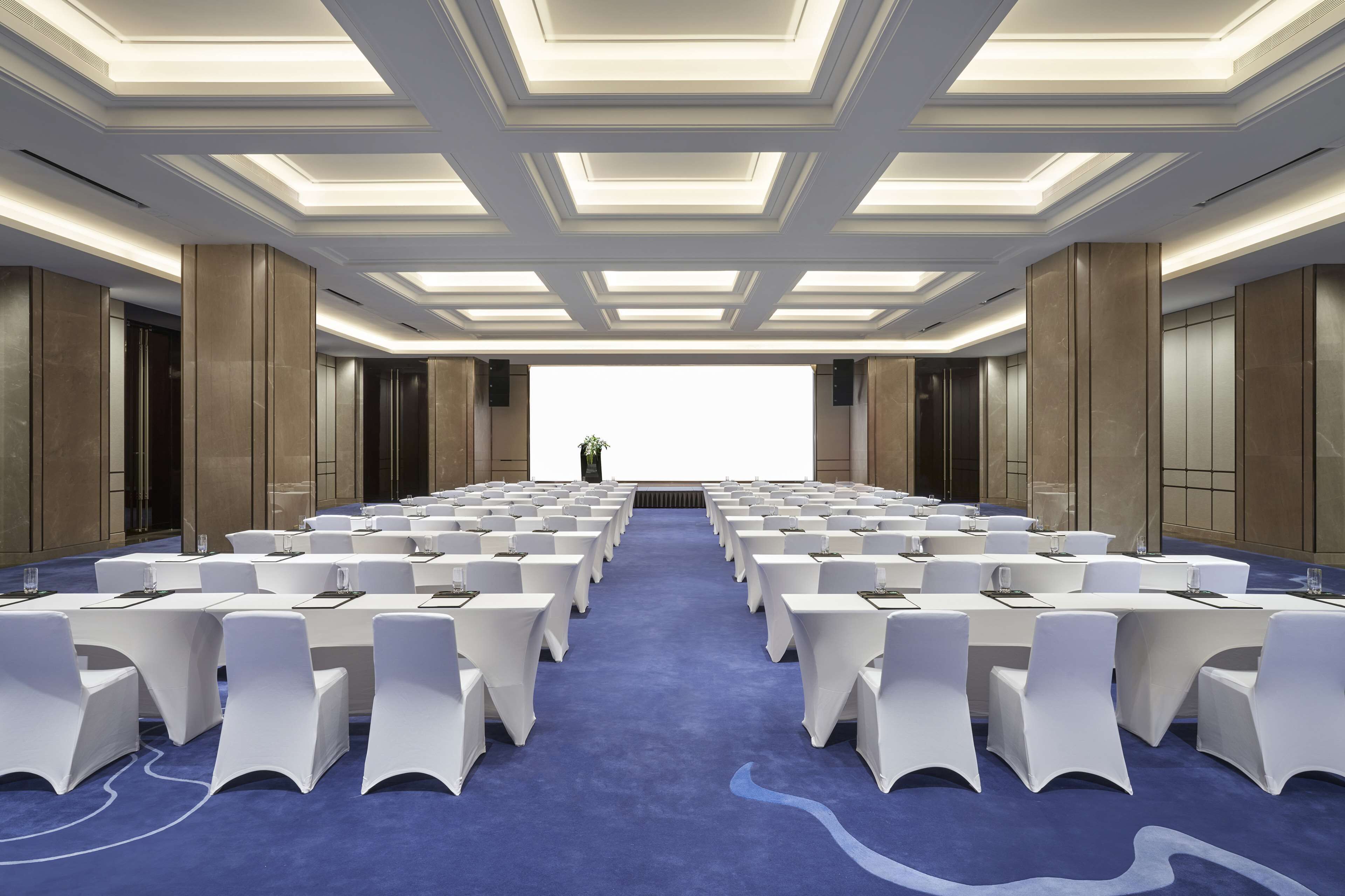 A conference room with rows of white chairs, a blue carpet, and a modern ceiling design.