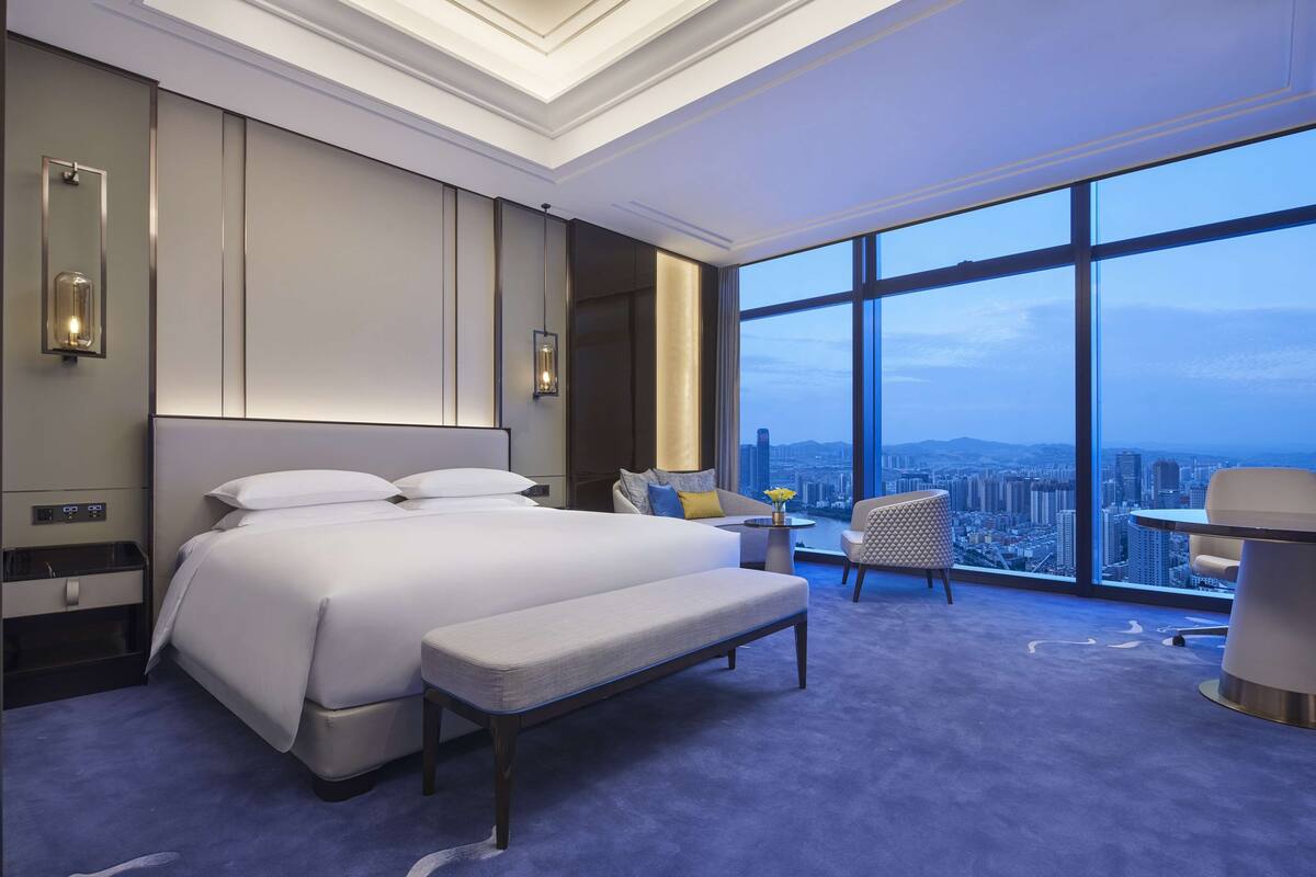 A modern hotel room with a large bed, a bench, a small table, and a chair. There is a large window offering a city view.