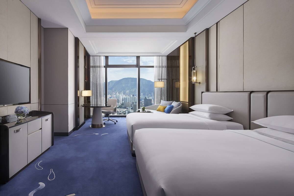 A hotel room with two beds, a desk, a chair, a TV, and a view of a cityscape through large windows.