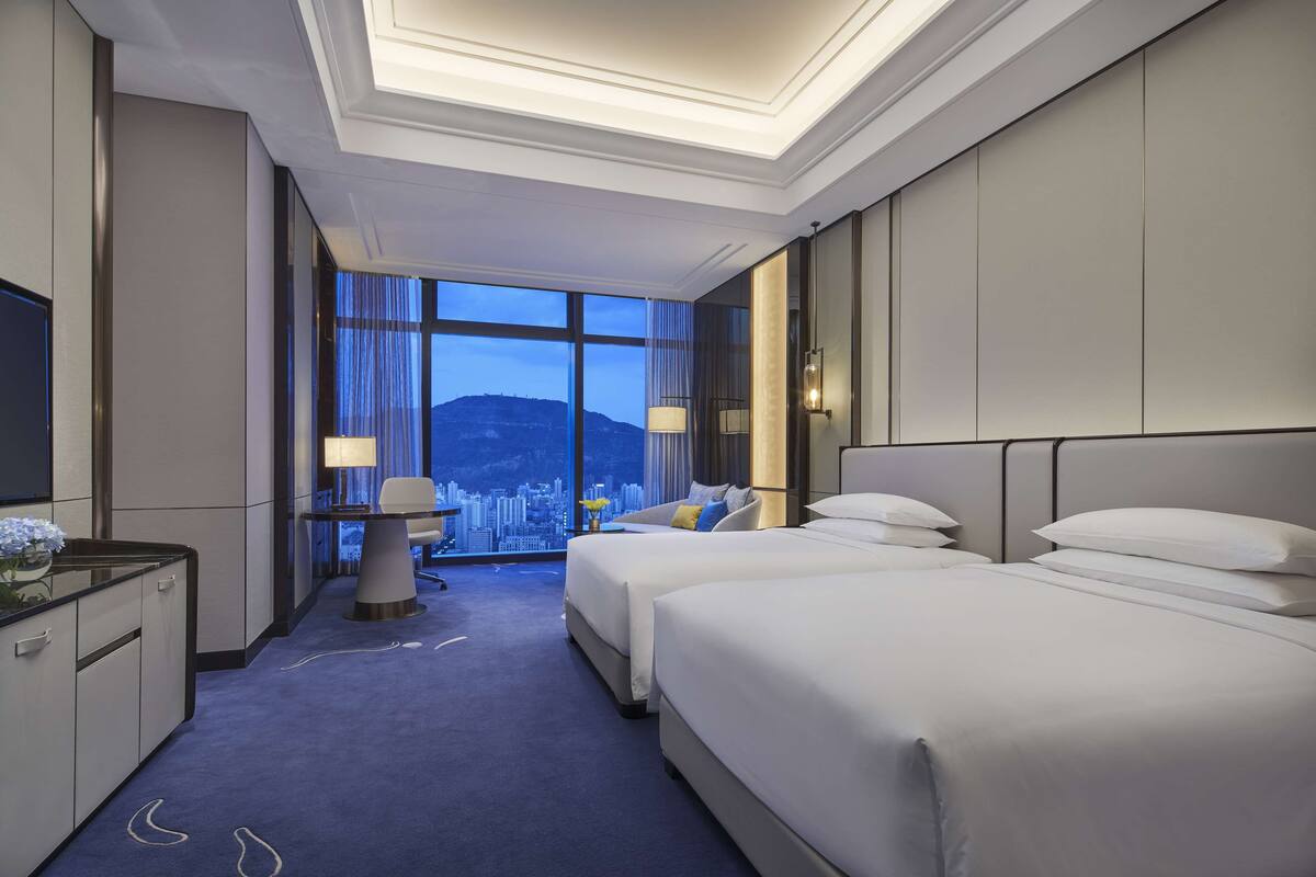 A hotel room with two beds, a desk, and a large window offering a city view.