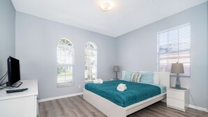 A bedroom with a bed, a nightstand, a lamp, a TV, and two windows with blinds.