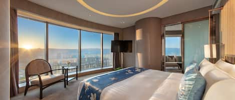 A modern hotel room with a large bed, a chair, a small table, a TV, and a city view.