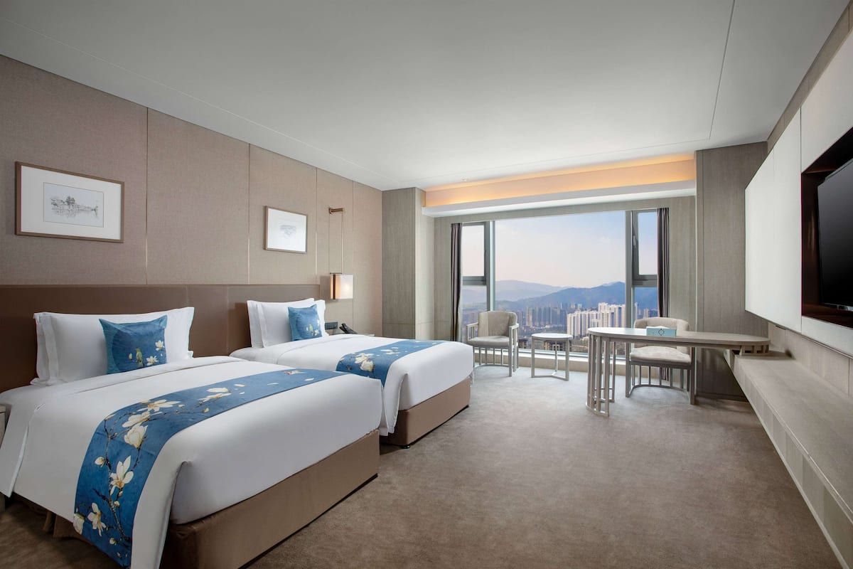 A hotel room with two beds, a desk, a chair, a TV, and a large window with a city view.