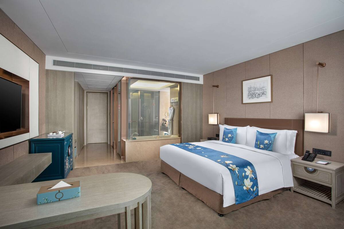 A hotel room with a bed, bedside tables, a desk, a TV, and a bathroom visible through an open door.
