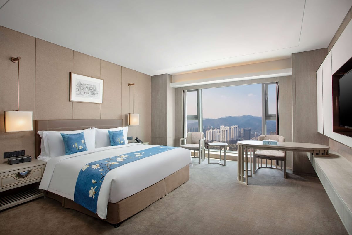 A modern hotel room with a large bed, bedside tables, a desk, and a view of a cityscape through large windows.