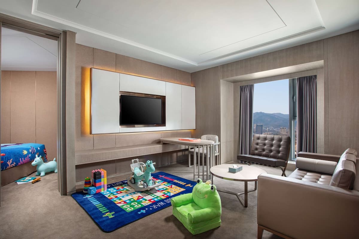 A modern hotel room with a flat-screen TV mounted on the wall, a plush green chair, a beige sofa, a small round table, and a play area with toys and a colorful rug.