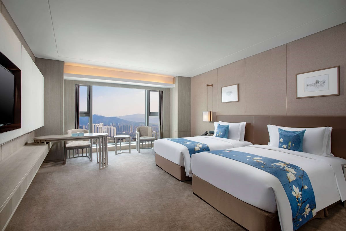 A hotel room with two beds, a desk, a chair, and a large window offering a city view.