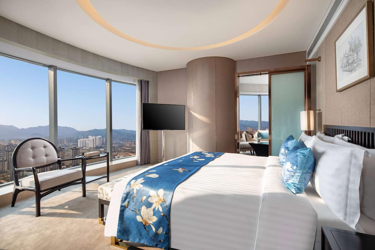 A modern hotel room with a large bed, a sitting area with a chair, a flat-screen TV, and a large window offering a city view.