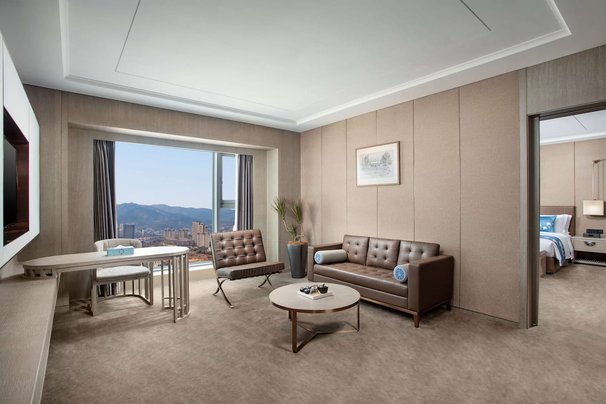 A modern hotel room with a large window, a sofa, a coffee table, and a TV.