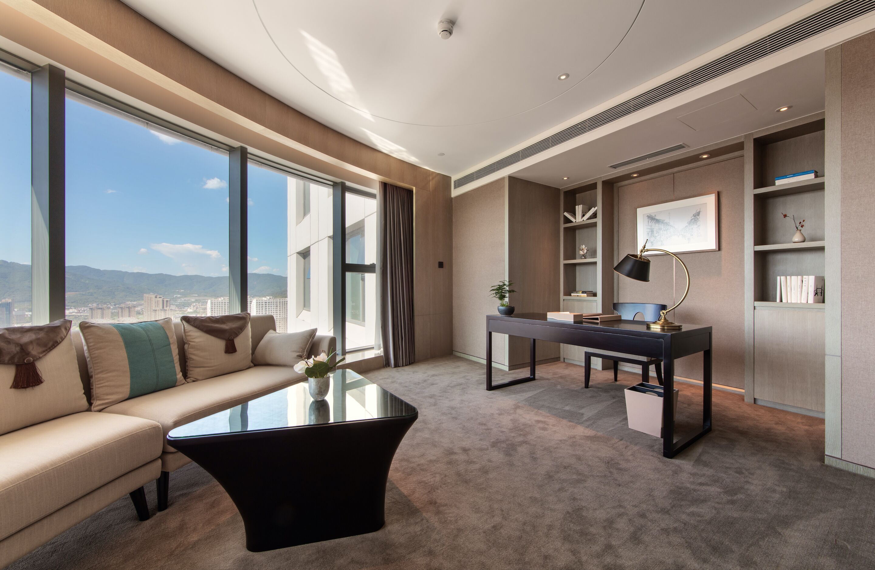 A modern hotel room with a sofa, a glass coffee table, a desk with a lamp, and a view of the city.