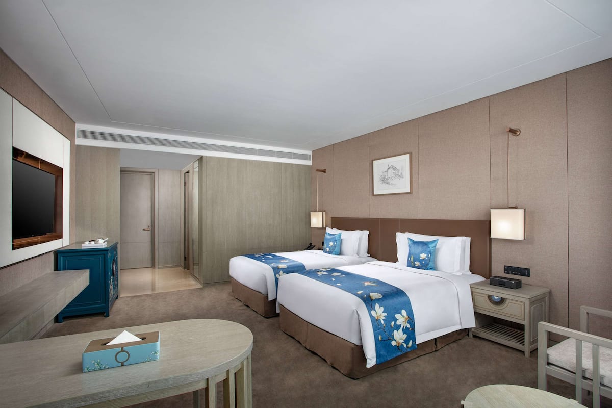A hotel room with two beds, a desk, a chair, a TV, and a bedside table.