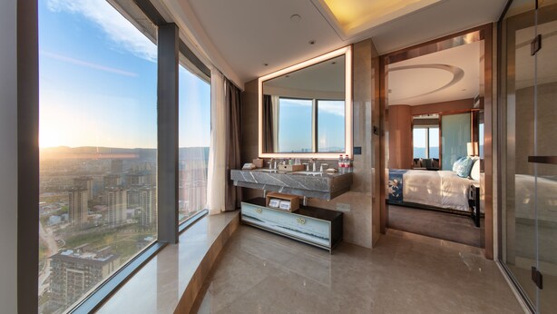 Une chambre d’hôtel moderne dotée d’une grande fenêtre, d’une salle de bain avec un meuble-lavabo en marbre et d’un lit offrant une vue sur la ville.