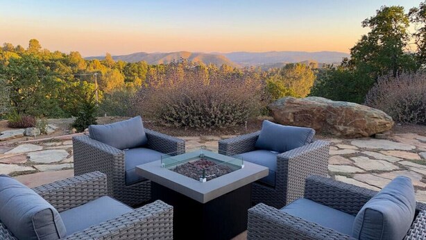 A patio with wicker furniture and a glass-top table, set against a backdrop of hills and trees.