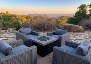 A patio with wicker furniture and a glass-top table, set against a backdrop of hills and trees. - Hot Tub, Privacy, Fire Pit, Pool Table, Ping Pong, Amazing Views! (Mariposa)