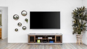 A living room with a flat-screen TV mounted on the wall, a wooden entertainment unit below, and a potted plant to the right.