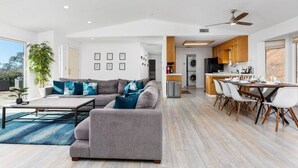 A modern living room with a gray sectional sofa, a dining area with a long table and chairs, and a kitchen with wooden cabinets and appliances.