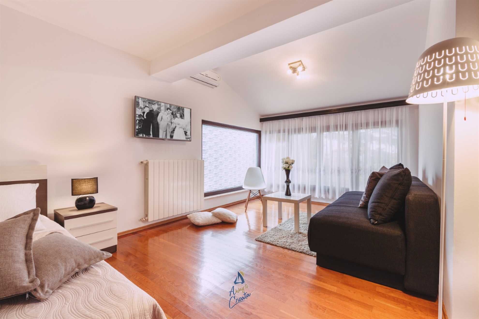A modern bedroom with a bed, a sofa, a small table, and a window with sheer curtains.