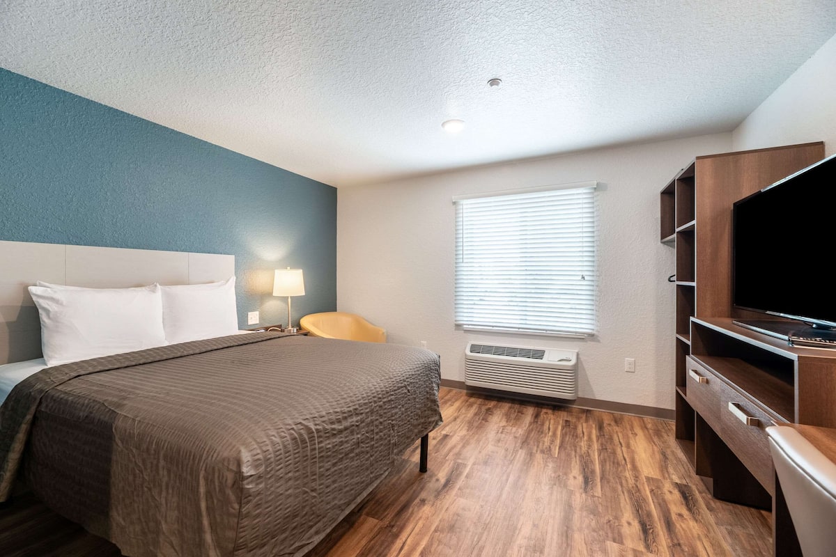 A hotel room with a bed, a desk, a TV, and a window with blinds.