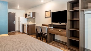 A compact hotel room with a kitchenette, a bed, a desk with a chair, and a TV. - WoodSpring Suites Sanford North I-4 Orlando Area (Sanford)