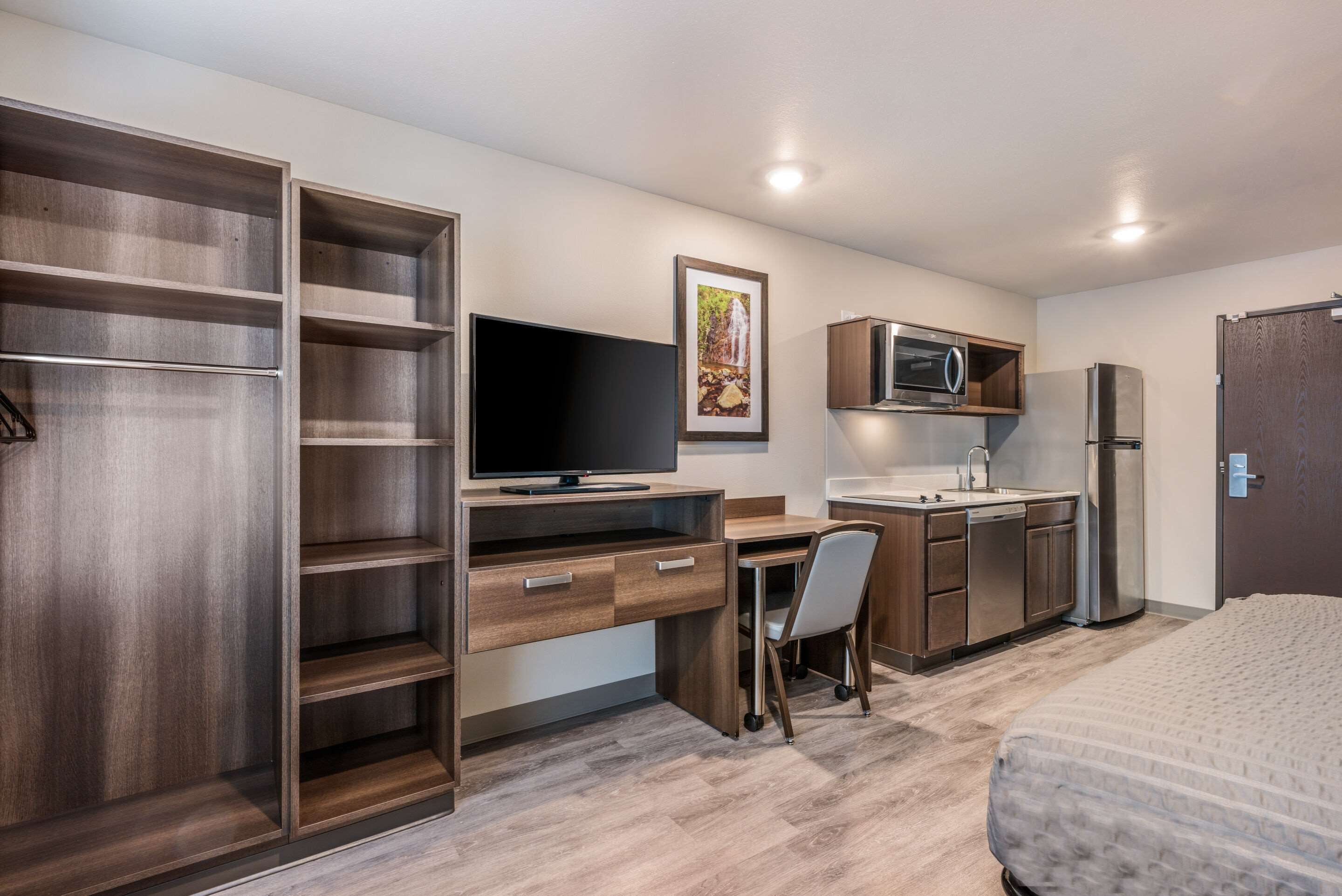 A compact hotel room with a wooden desk, a television, a kitchenette, and a bed.