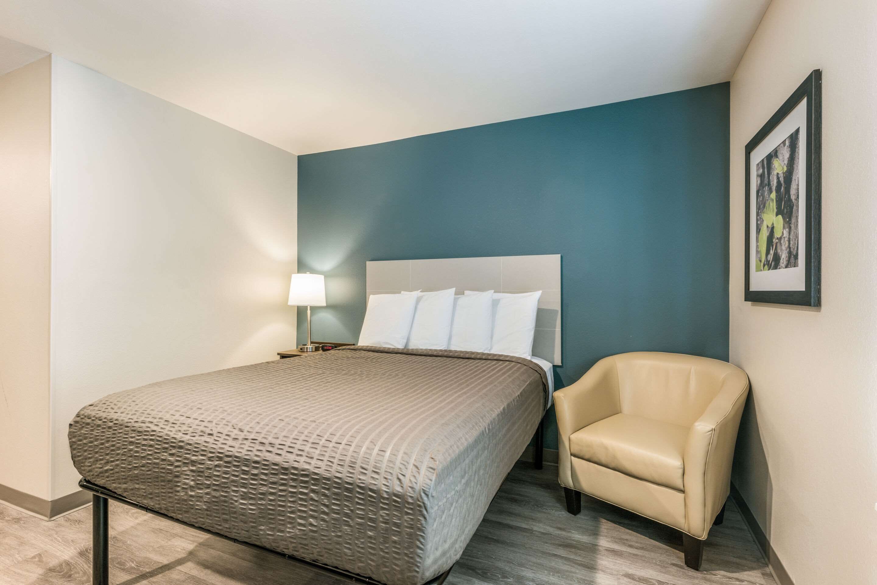 A hotel room with a bed, a chair, a nightstand, and a wall-mounted lamp.