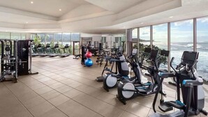 Ein großzügiges Fitnessstudio mit Laufbändern, Hanteln und Trainingsbällen, mit großen Fenstern und Blick auf den Ozean.