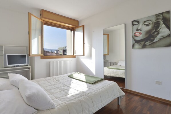 A bedroom with a bed, a TV, a window, and a painting on the wall.
