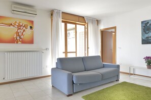 A living room with a grey sofa, a green rug, a radiator, a painting, and a door leading to another room.