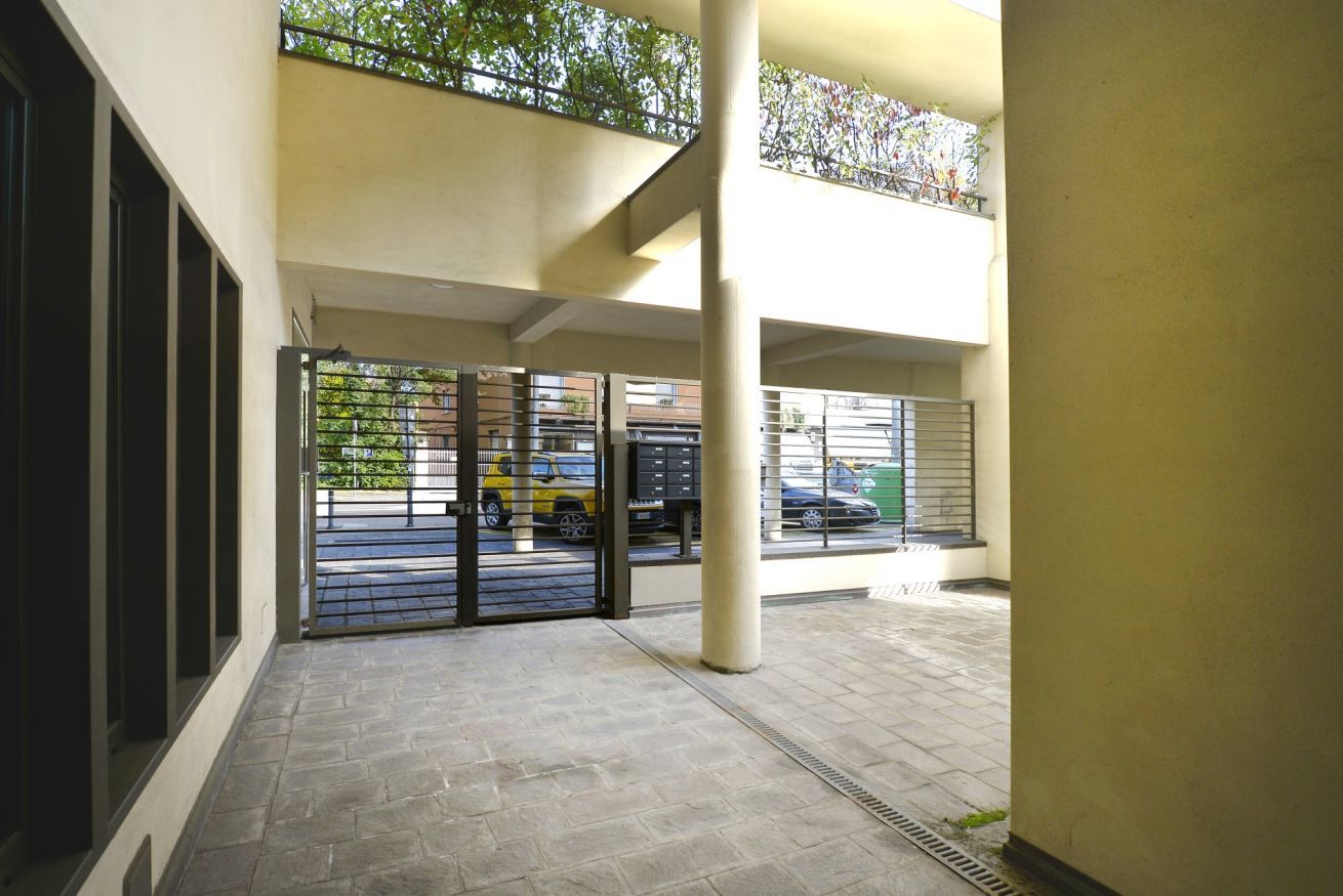 Un'area esterna coperta con pavimento piastrellato, un cancello in metallo e vista su auto parcheggiate attraverso il cancello.