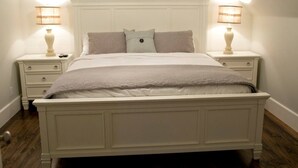 Een netjes opgemaakt bed met een wit hoofdeinde, twee nachtkastjes met lampen, en een raam met zonwering.