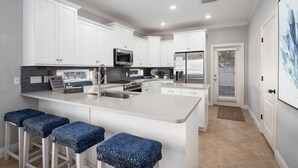 Cocina moderna con armarios blancos, una isla central con taburetes de bar, electrodomésticos de acero inoxidable y un amplio ventanal con persianas.