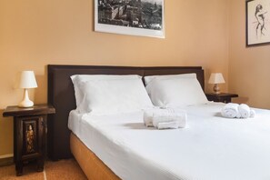 A neatly made bed with white linens and pillows, two bedside tables with lamps, a framed cityscape picture on the wall, and a small statue on one of the tables.
