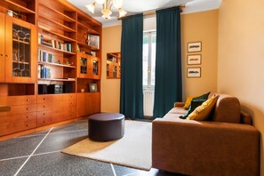 A cozy living room with a brown sofa, a wooden bookshelf filled with books and decorative items, a small round ottoman, and a large window with curtains.