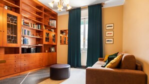 A cozy living room with a brown sofa, a wooden bookshelf filled with books and decorative items, a small round ottoman, and a large window with curtains. - UR-NEST Lame, Spacious one bedroom (Bologna)