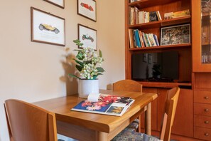 A wooden dining table with chairs, a vase with flowers, and a book. There is a bookshelf with books and framed pictures on the wall.