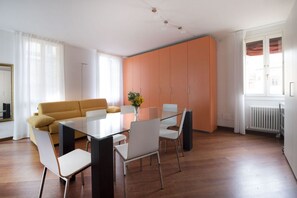 A modern living room with a glass dining table, white chairs, a mustard-colored sofa, and orange cabinets. - UR-NEST Moline, Lovely three bedroom two bathroom apartment (Bologna)