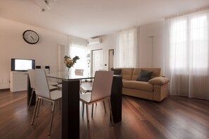 A modern living room with a sofa, a dining table with chairs, a TV, and a clock on the wall.