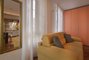 A living room with a yellow sofa, a large mirror, and a kitchen visible through the window.