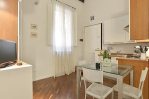 A compact kitchen with white cabinets, a glass dining table, and a TV on a stand.
