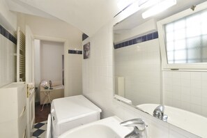 Une salle de bain avec un lavabo blanc, un miroir et une baignoire blanche. Une porte donne accès à une autre pièce.