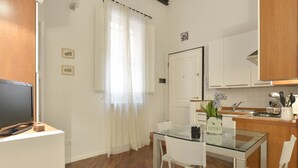 Una cocina compacta con armarios blancos, una mesa de comedor de vidrio y un televisor sobre un mueble.